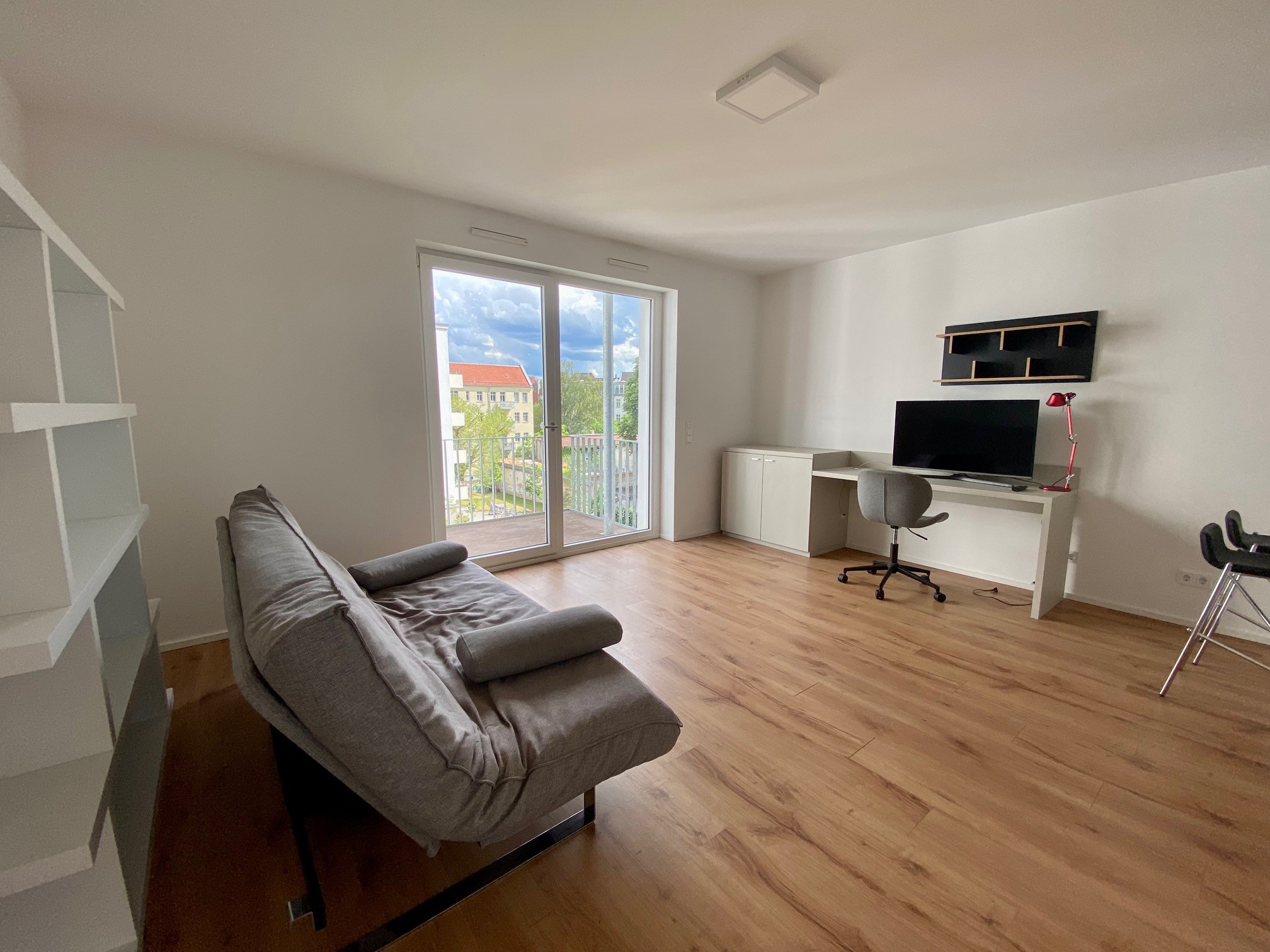 Helles, teileingerichtetes Wohnzimmer mit warmem Laminatboden, frisch gestrichenen weißen Wänden und einer großen Fensterfront mit Zugang zu einem Balkon. Der Raum verfügt über eine funktionale Grundausstattung mit einem grauen Sofa, einem integrierten Home-Office-Bereich mit Schreibtisch und Bürostuhl sowie einem weißen Regal. Dieses authentische Immobilienfoto eignet sich ideal für Immobilien-Exposés, zur Visualisierung des Raumkonzepts oder als Grundlage für eine digitale Aufwertung durch virtuelles Home Staging.