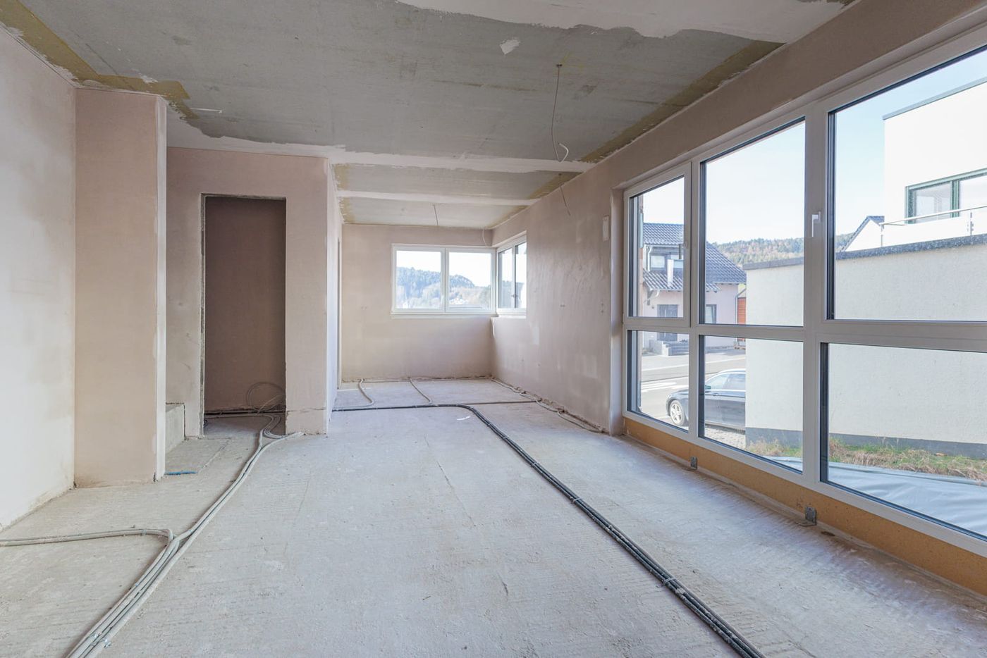 Vorher-Bild für virtuelles Home Staging: Ein heller, langgestreckter Raum im Rohbauzustand. Die Baustelle zeigt einen nackten Betonboden mit freiliegenden Leitungen, unverputzte Wände und eine unfertige Decke. Große, moderne Fensterfronten sorgen für viel Tageslicht und bieten ideales Potenzial für die digitale Visualisierung eines offenen Essbereichs.