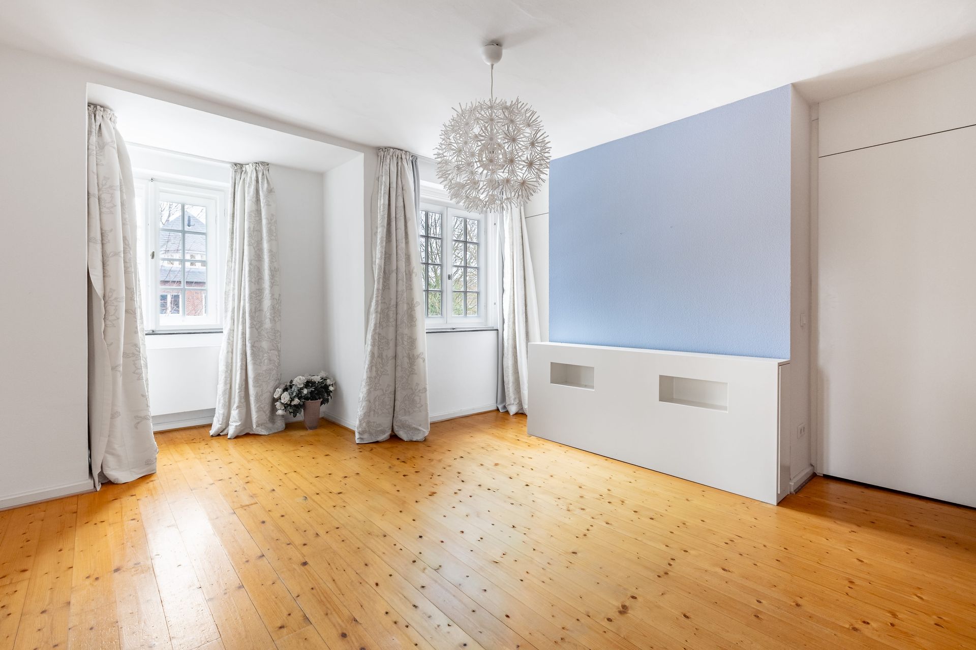 Vorher-Bild für virtuelles Home Staging: Ein leeres, unmöbliertes Schlafzimmer mit einem hellen, rustikalen Dielenboden. Der Raum verfügt über zwei Fenster mit hellen Vorhängen, eine auffällige kugelförmige Deckenleuchte und eine hellblaue Akzentwand mit einer weißen, halbhohen Vorwandinstallation. Dieser leere Zustand bietet die ideale Ausgangsbasis für eine fotorealistische digitale Möblierung, um potenziellen Käufern bei der Immobilienvermarktung die optimale Raumnutzung und das volle Potenzial zu veranschaulichen.