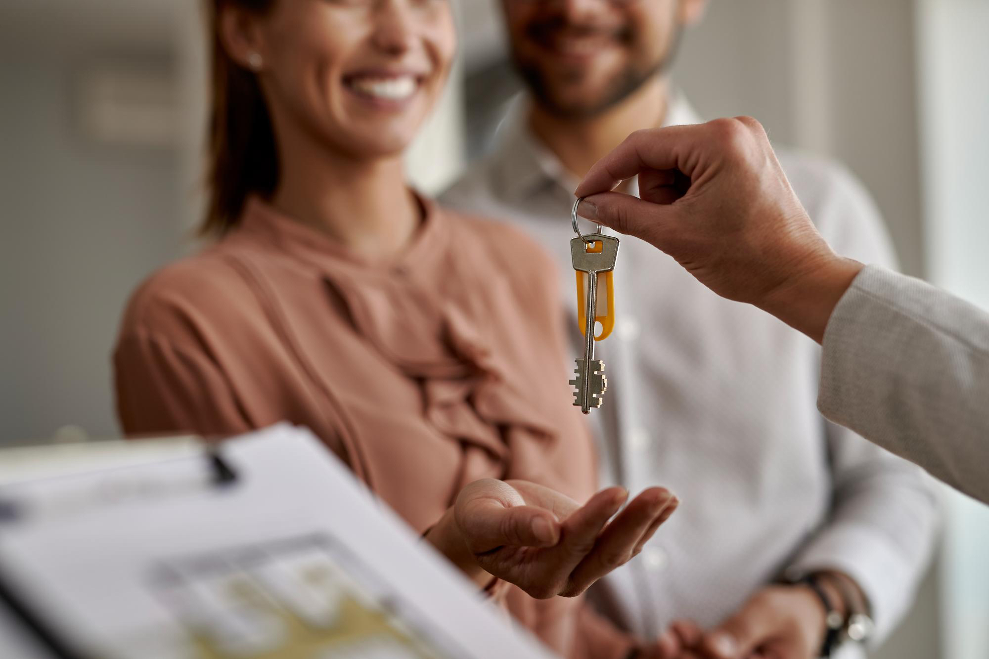 Ein Immobilienmakler überreicht den Schlüsselbund mit einem gelben Anhänger an eine lächelnde Käuferin, die ihn entgegennimmt, während ihr Partner im Hintergrund steht. Ein unscharfer Grundriss ist im Vordergrund sichtbar.